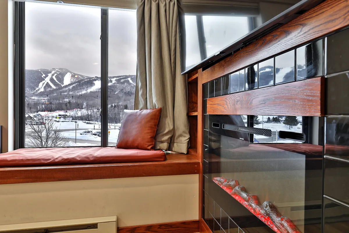 This image features a cozy interior scene with a prominent fireplace, a cushioned bench positioned by a large window, and inviting seating. Through the window, you catch a glimpse of the outdoors, suggesting a tranquil mountain setting perfect for both relaxation and inspiration.