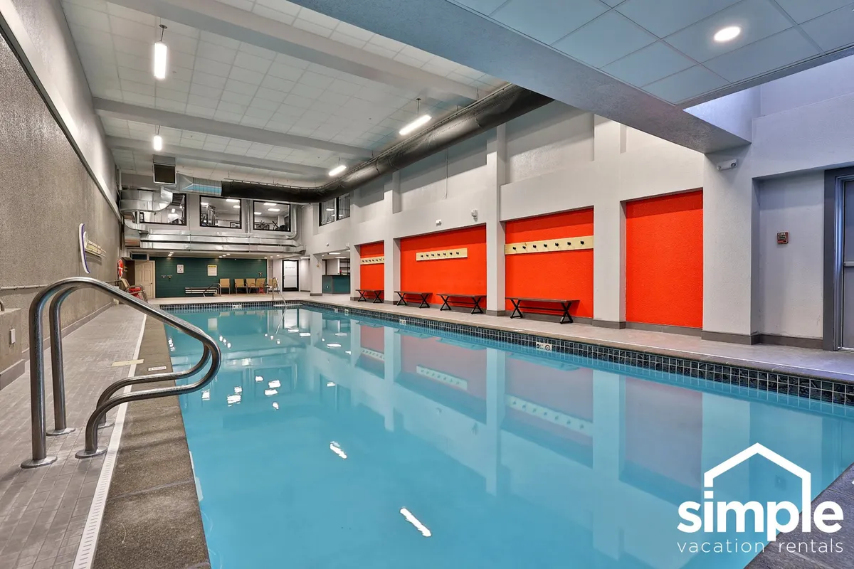The image shows a vibrant indoor swimming pool area with red accent walls, a seating area, and ample space for relaxing and socializing by the water. The mood is relaxed and fun, inviting guests to enjoy activities such as swimming or unwinding after a long day on the slopes.