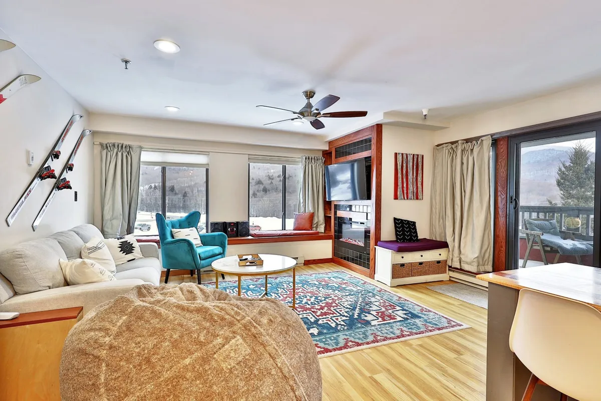 The image showcases a stylish and inviting living room featuring a plush sofa, two vibrant turquoise chairs, a round coffee table, and a cozy fireplace. Expansive windows frame breathtaking mountain views, flooding the space with natural light and creating a warm, welcoming atmosphere ideal for relaxing or gathering after a day on the slopes.