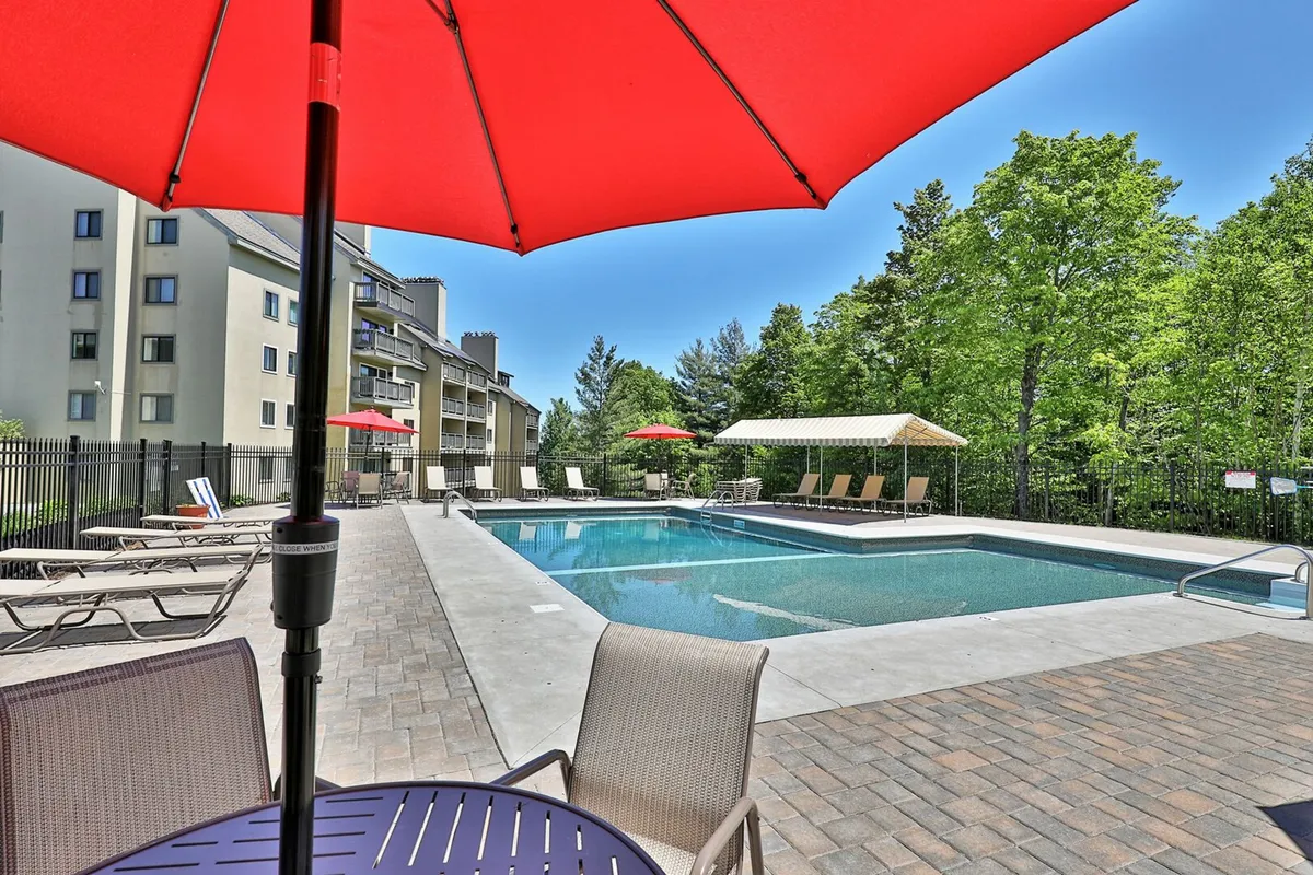 An exterior view of a sparkling outdoor swimming pool surrounded by red umbrellas, lounge chairs, and lush green trees, creating a leisurely and refreshing summer scene. The area is ideal for social gatherings, sunbathing, and poolside barbecues.