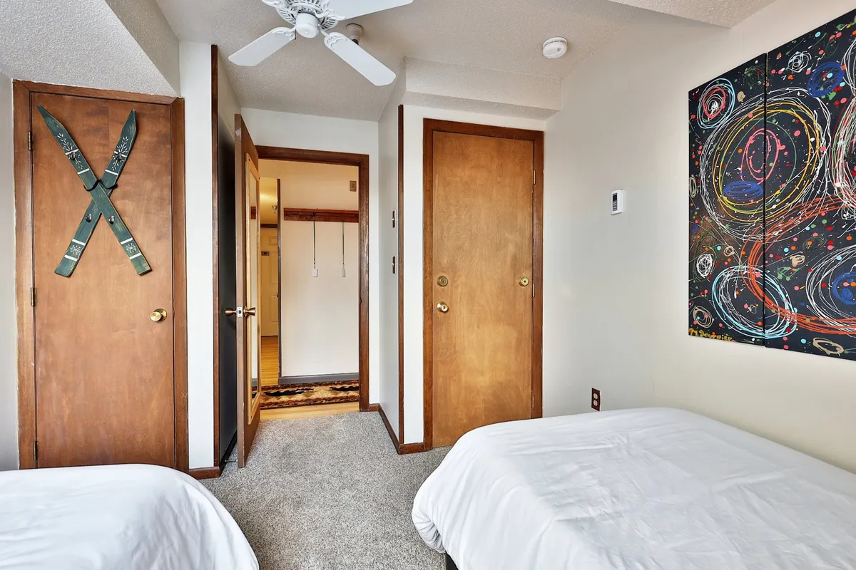 A cozy second bedroom featuring two twin beds with crisp white bedding, cheerful abstract wall art, and ski-themed decor. The room is inviting, with ample space for siblings or friends.