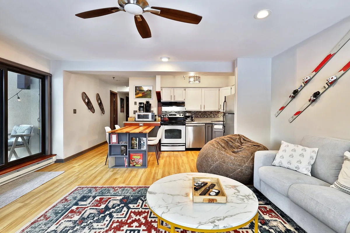 A welcoming space with a coffee table and decorative tray, bean bag chair, open kitchen area, and ski-themed decorations on the wall. The scene is lively and casual, emphasizing both relaxation and the social aspect of sharing meals or movie nights.