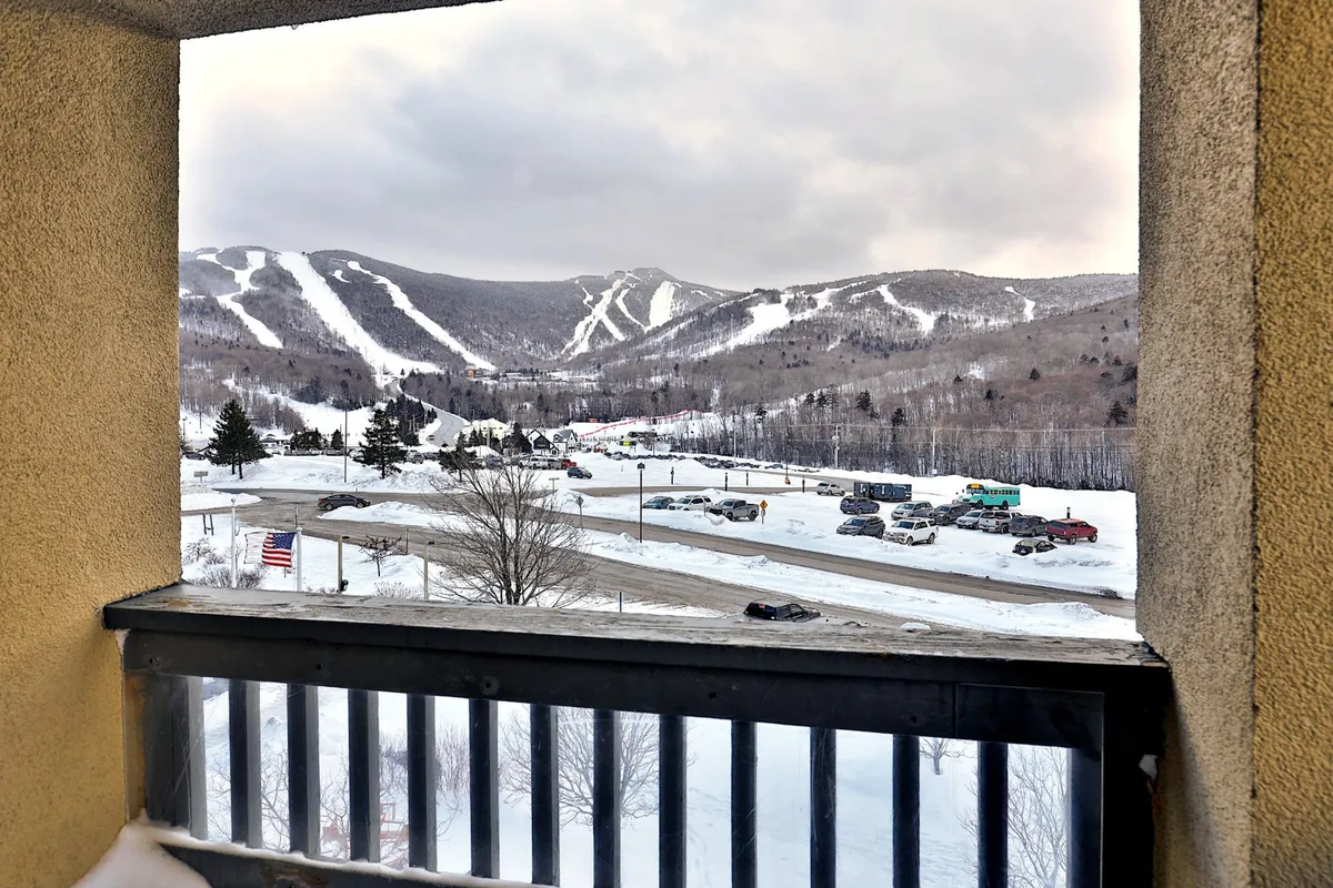 A breathtaking view of snow-covered mountains and ski slopes, with the American flag and a parking lot in the foreground. The serene scene showcases skiers and snowboarders in the distance, encapsulating the thrill and natural beauty of the Killington area during ski season.