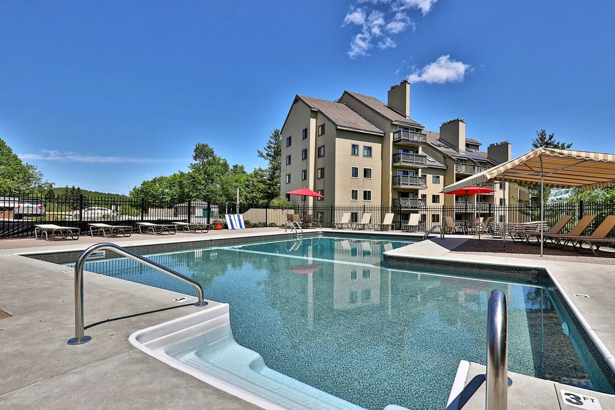 This image showcases an inviting outdoor swimming pool surrounded by lounge chairs, a canopy, and umbrellas, with the resort building visible in the background. The scene is leisurely and welcoming, suggesting summertime relaxation and family fun in the sun.