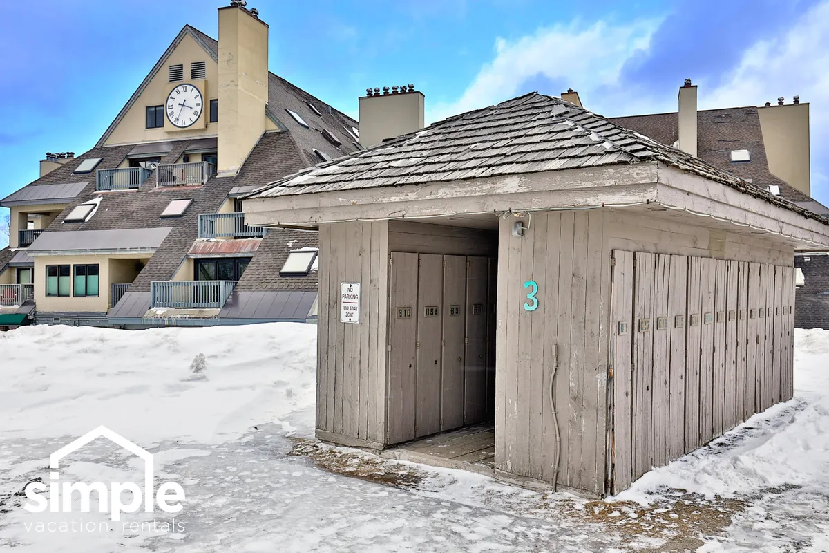 This exterior image features the resort's storage locker building and main building with a clock tower, all set against a snow-covered landscape. There are visible pathways and communal spaces where guests gather before heading out for outdoor activities.