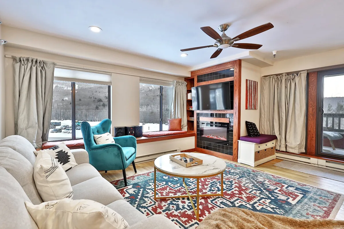A cozy living room area featuring a blue accent chair and a round coffee table, set near a modern electric fireplace. Large windows frame sweeping mountain views, and the arrangement includes ample comfortable seating, making it an inviting place for relaxation or socializing.